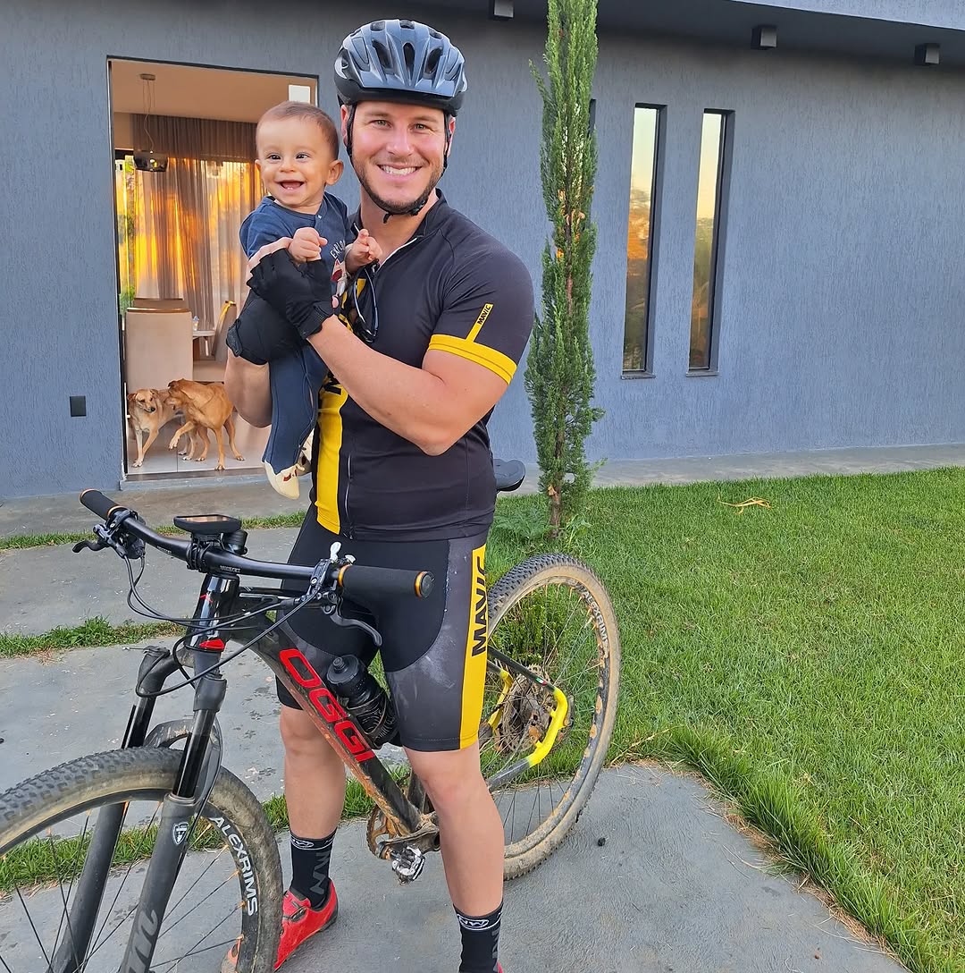 Felipe Malafaia segurando seu filho no colo após uma pedalada de mountain bike, com a bicicleta ao lado em frente à sua casa